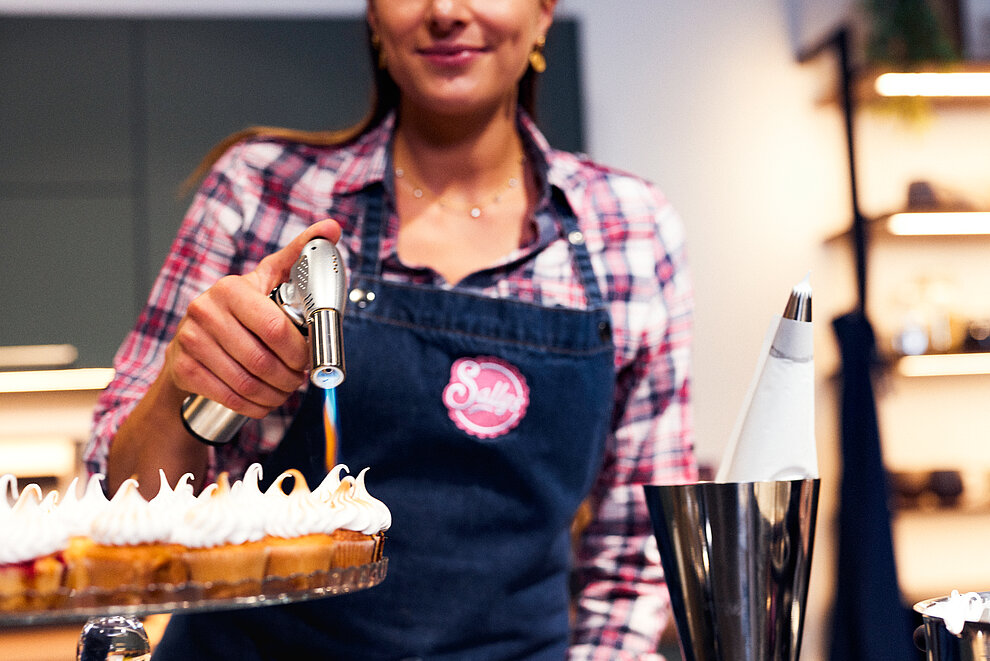 A baker uses a culinary torch to caramelize the meringue topping on a pie, wearing an apron with a cheerful logo at a professional kitchen.