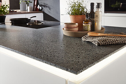 Elegante Küchenarbeitsplatte mit raffiniertem Terrazzo-Design, auf der ordentlich angeordnete Küchenaccessoires und grüne Topfkräuter den modernen Kochbereich verschönern.