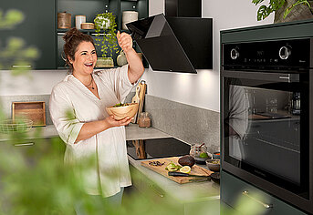 A smiling person seasoning her food in a modern kitchen with stylish black appliances and greenery in the background.