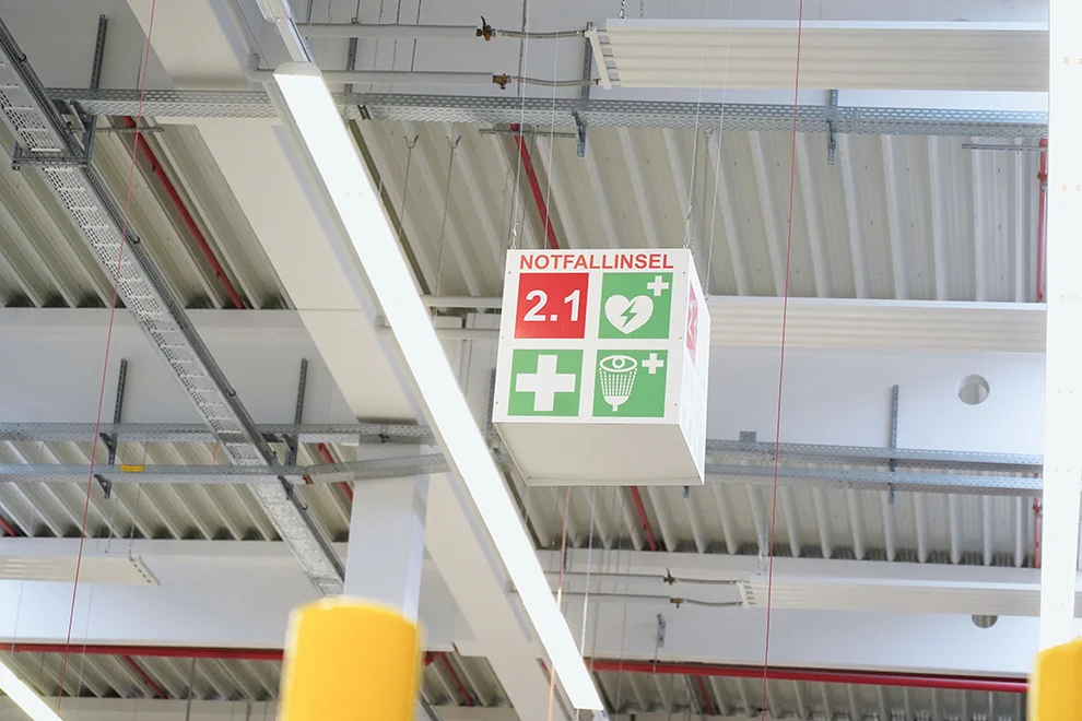 An emergency first aid station, marked with a green cross and signage, hangs from a ceiling with exposed beams and infrastructure in an industrial setting.