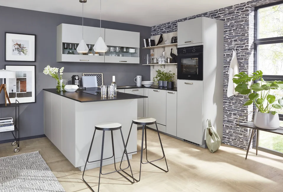 Cuisine en U compacte avec façade TOUCH 341 en gris pierre supermate, conçue de manière fonctionnelle pour les petits espaces, armoires suspendues modernes avec façade en verre et îlot stylé avec possibilité de s'asseoir.