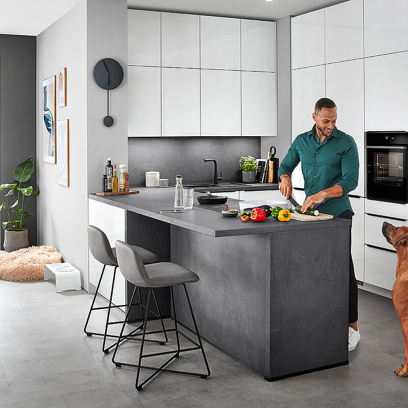 Stylowa nowoczesna scena kuchenna z mężczyzną przygotowującym jedzenie na wyspie kuchennej, przyglądającym się pieskiem, odzwierciedlająca wygodny, współczesny styl życia w domu.