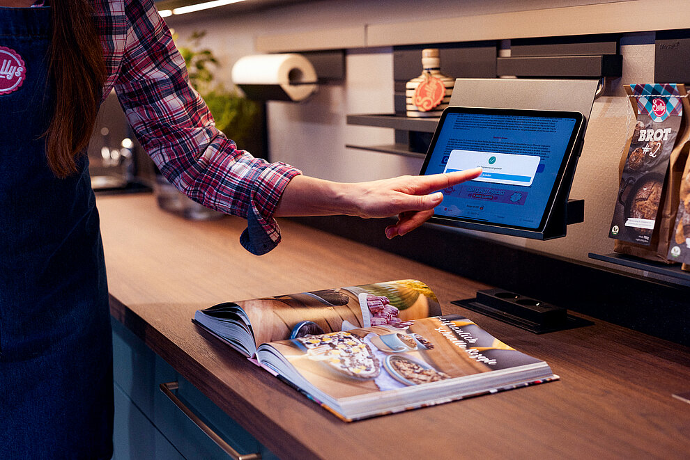 Eine Person interagiert mit einem Touchscreen-Gerät, das auf einem Tresen einen Anmeldebildschirm anzeigt, mit einem Kochbuch und verpackten Lebensmittelprodukten in der Nähe.