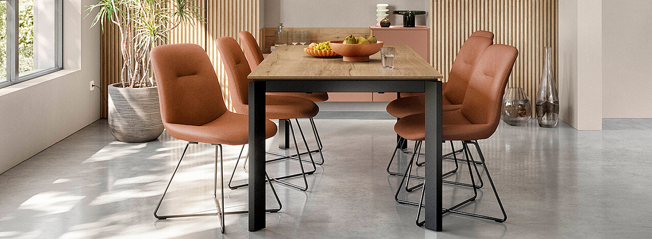 Moderne eetkamer met een houten tafel met zwarte poten, omringd door comfortabele stoelen op een gepolijste betonnen vloer tegen een geribbelde muurachtergrond.