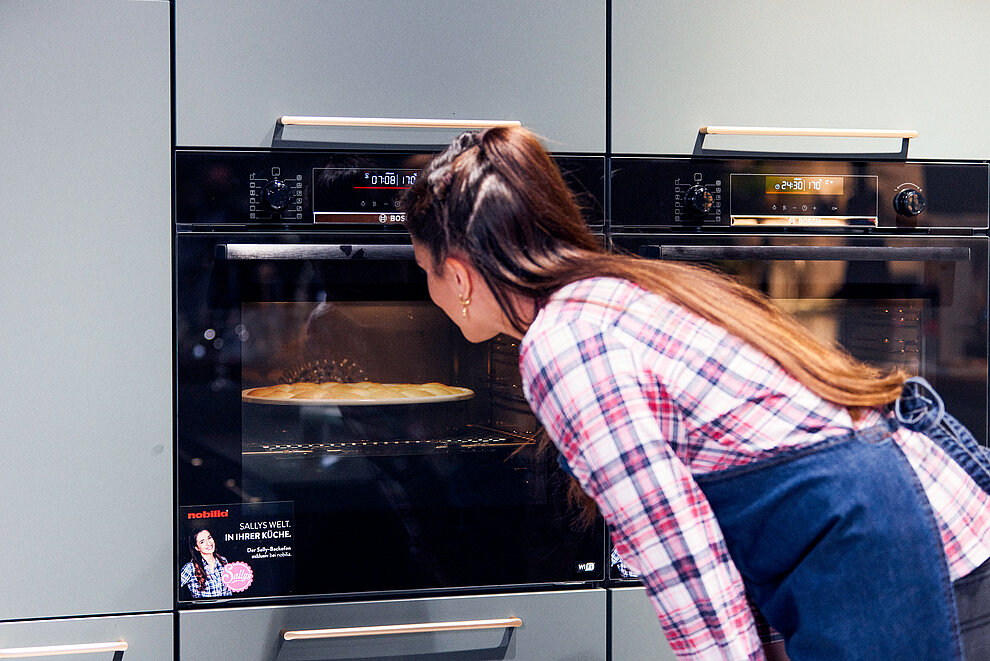 Een persoon gluurt in een oven, kijkend naar een taart terwijl deze bakt, in een moderne keuken met strakke apparaten en een strak ontworpen interieur.