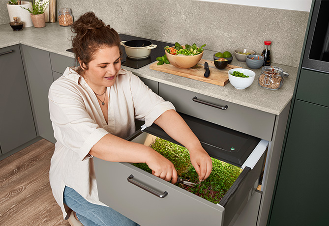 Una mujer sonríe mientras cuida hierbas en un jardín de cajón de cocina moderno, rodeada de ingredientes frescos y utensilios de cocina.