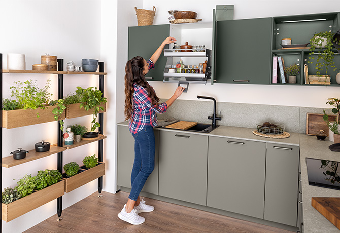 Donna in una cucina moderna con armadi verdi che raggiunge gli oggetti circondata da erbe fresche e mensole in legno, raffigurando uno stile di vita eco-friendly.