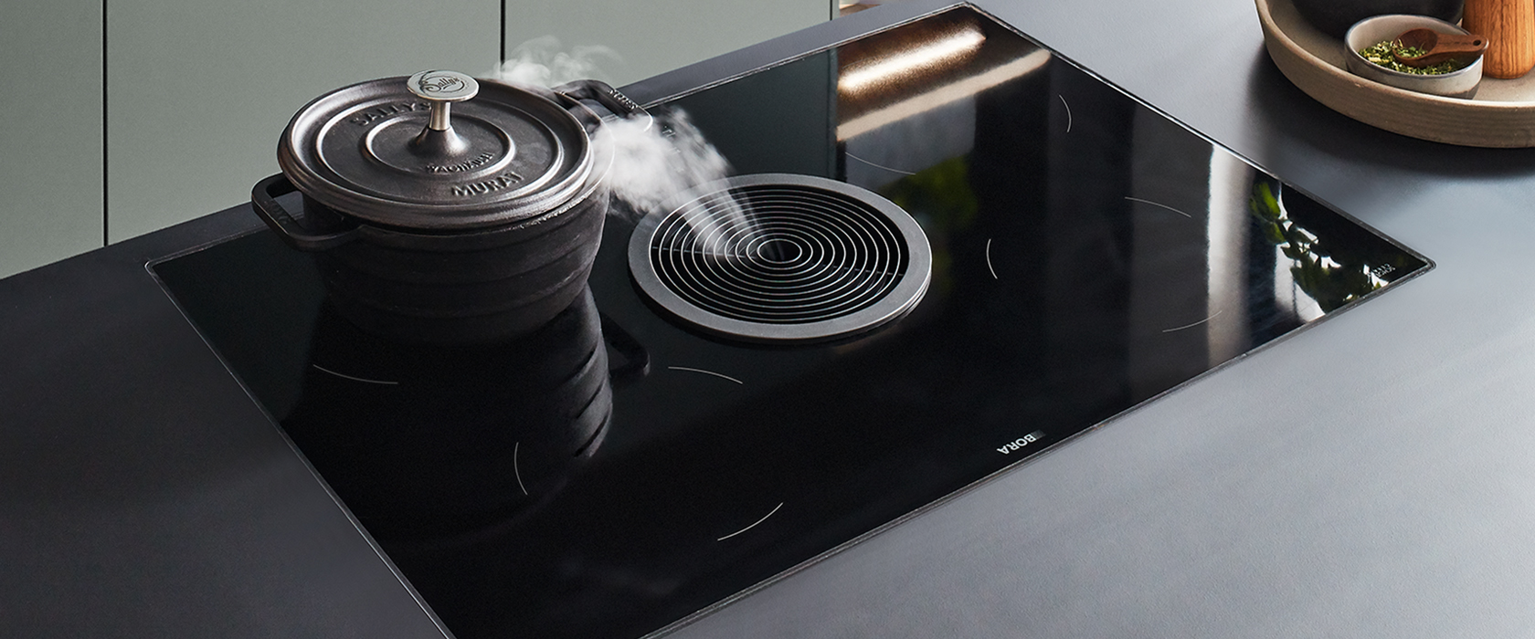 Strakke, moderne inductiekookplaat met een actieve kookpot, waar stoom uit opstijgt, geplaatst in een eigentijdse keuken met subtiele aardetinten.