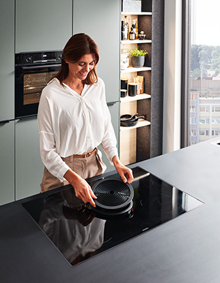 Femme dans une cuisine moderne utilisant un système d'extraction intégré à aspiration descendante tout en se tenant à côté d'une table de cuisson à induction, mettant en valeur un mélange de technologie et de design élégant.
