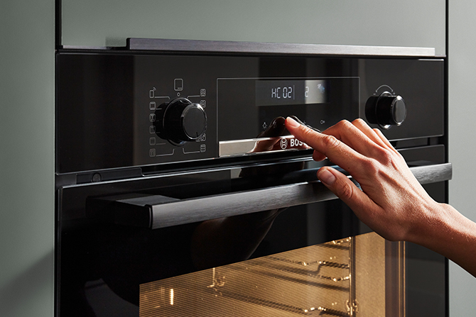 A person's hand adjusting the modern, sleek controls on a built-in black oven with a clear digital display, in a contemporary kitchen setting.
