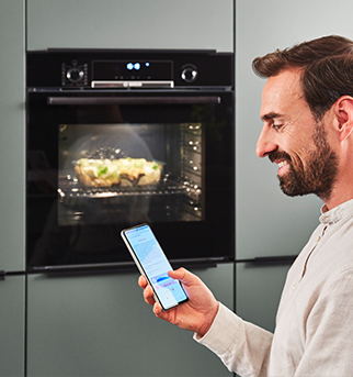 Uomo con la barba che utilizza uno smartphone per controllare un forno intelligente con una chiara vista del cibo che viene cucinato all'interno.