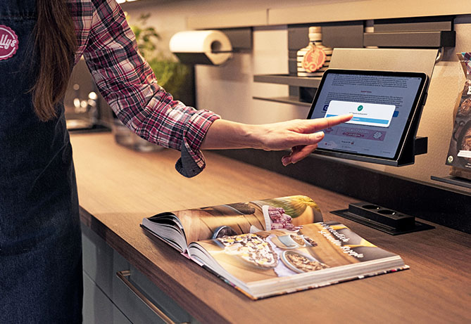 Persona usando una tableta digital montada en la encimera de la cocina para navegar por un sitio web, con un libro de cocina e ingredientes cerca, sugiriendo una actividad de cocina.