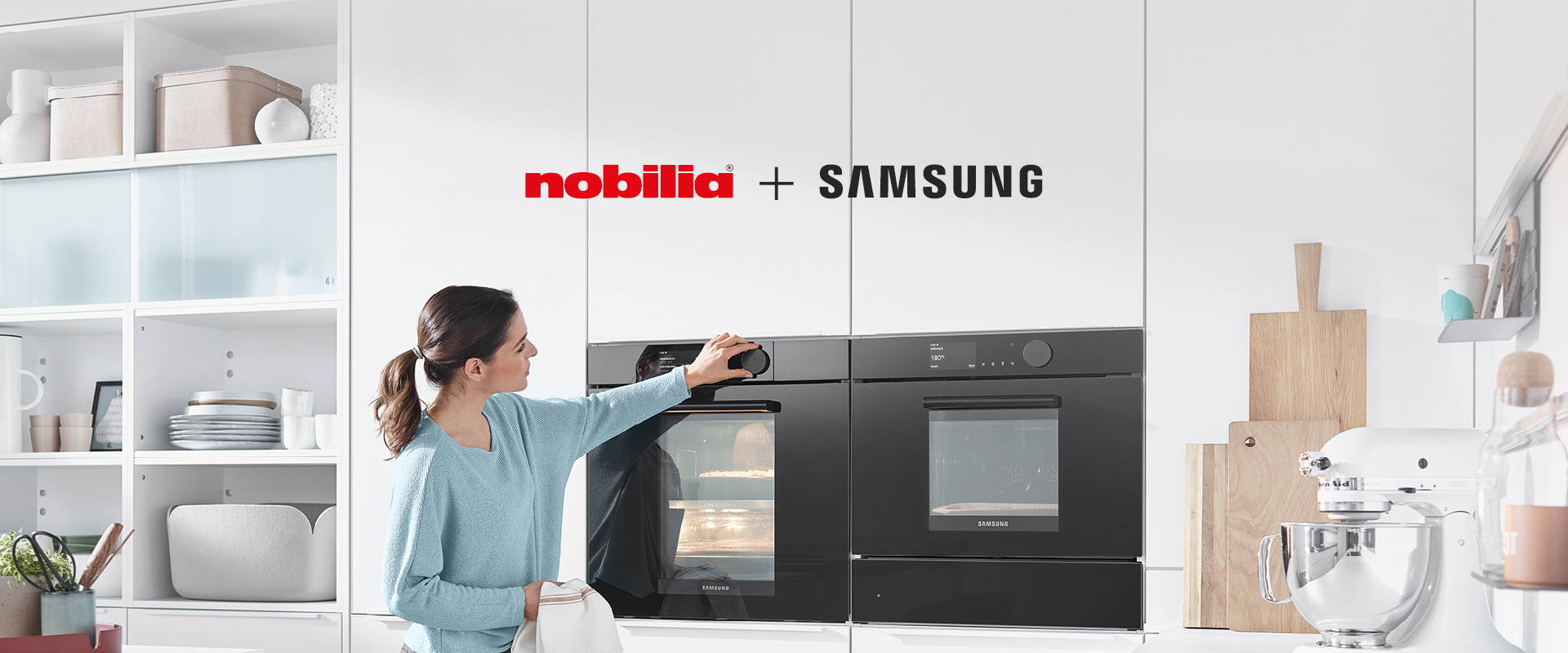 Moderne Kücheneinstellung, die eine Frau zeigt, die mit einem hochmodernen Samsung-Ofen interagiert, der in elegante, weiße Nobilia-Schränke eingebaut ist.