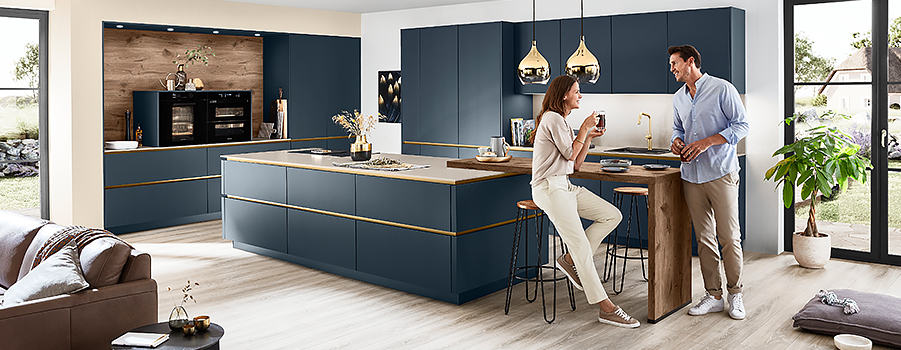 Moderní kuchyně s elegantní modrou kuchyňskou linkou a zlatými doplňky, kde dva lidé užívají teplý rozhovor při pití v stylovém, útulném prostoru.