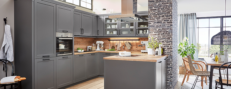 Spacious modern kitchen with gray cabinetry, wooden countertops, stainless steel appliances, and a stylish stone accent wall, complemented by natural light and greenery.