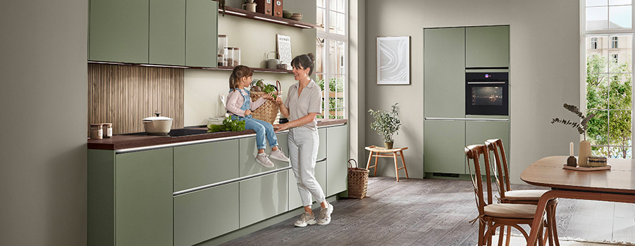 Diseño de cocina moderna con gabinetes blancos limpios, una isla central, y una persona cocinando en un espacio luminoso y aireado con vista a la ciudad a través de grandes ventanas.