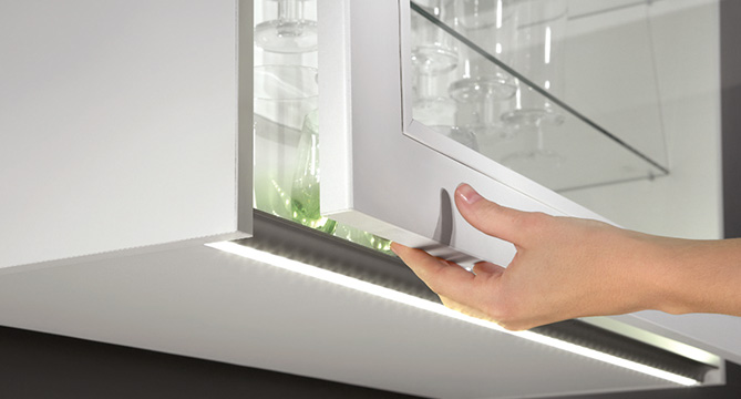 Een hand van een persoon opent een moderne witte kast met glazen planken, verlicht door onder-kastverlichting in een strakke keukenomgeving.