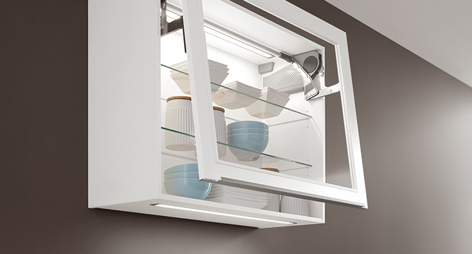 Armario de cocina moderno montado en la pared con estantes de vidrio, abierto para mostrar platos y tazas ordenados, contra una pared de tonos cálidos para un diseño minimalista de hogar.