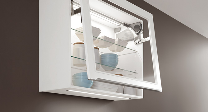 Modern wall-mounted kitchen cabinet with glass shelves, opened to reveal neatly arranged dishes and cups, against a warm-toned wall for a minimalist home design.