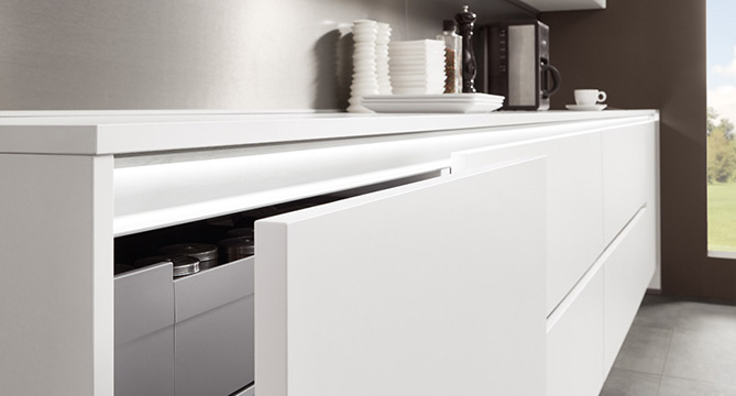 Cuisine moderne avec des tiroirs blancs lisses partiellement ouverts, révélant des ustensiles organisés, aux côtés d'une machine à café et d'assiettes sur le comptoir.