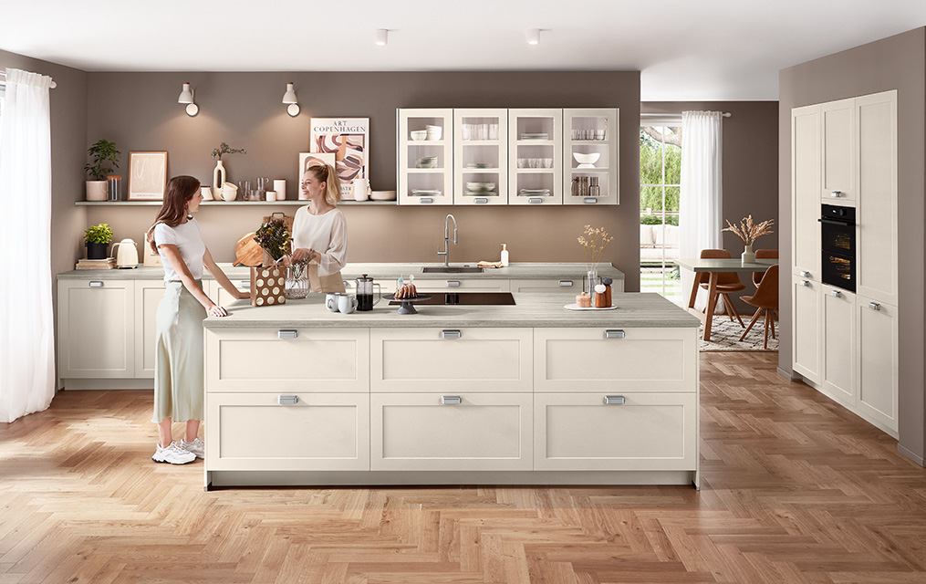 Moderne Landhausküche in Sand matt mit Front Nordic 788, Kochinsel, klarer Linienführung und heller Travertin-Arbeitsplatte vor warmem Wandton.