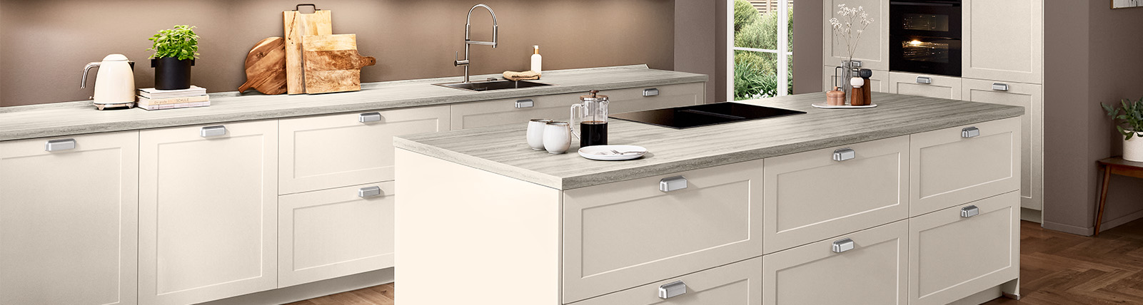 Moderne Landhausküche in Sand matt mit Front Nordic 788, Kochinsel, klarer Linienführung und heller Travertin-Arbeitsplatte vor warmem Wandton.