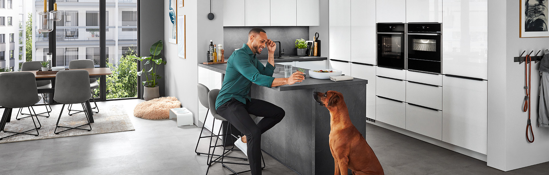 Moderne Küche mit Front Novalux 516 in Alpinweiß Hochglanz, elegante Kochinsel in Steinoptik, entspannte Wohnatmosphäre mit Essbereich und Hund im Vordergrund.