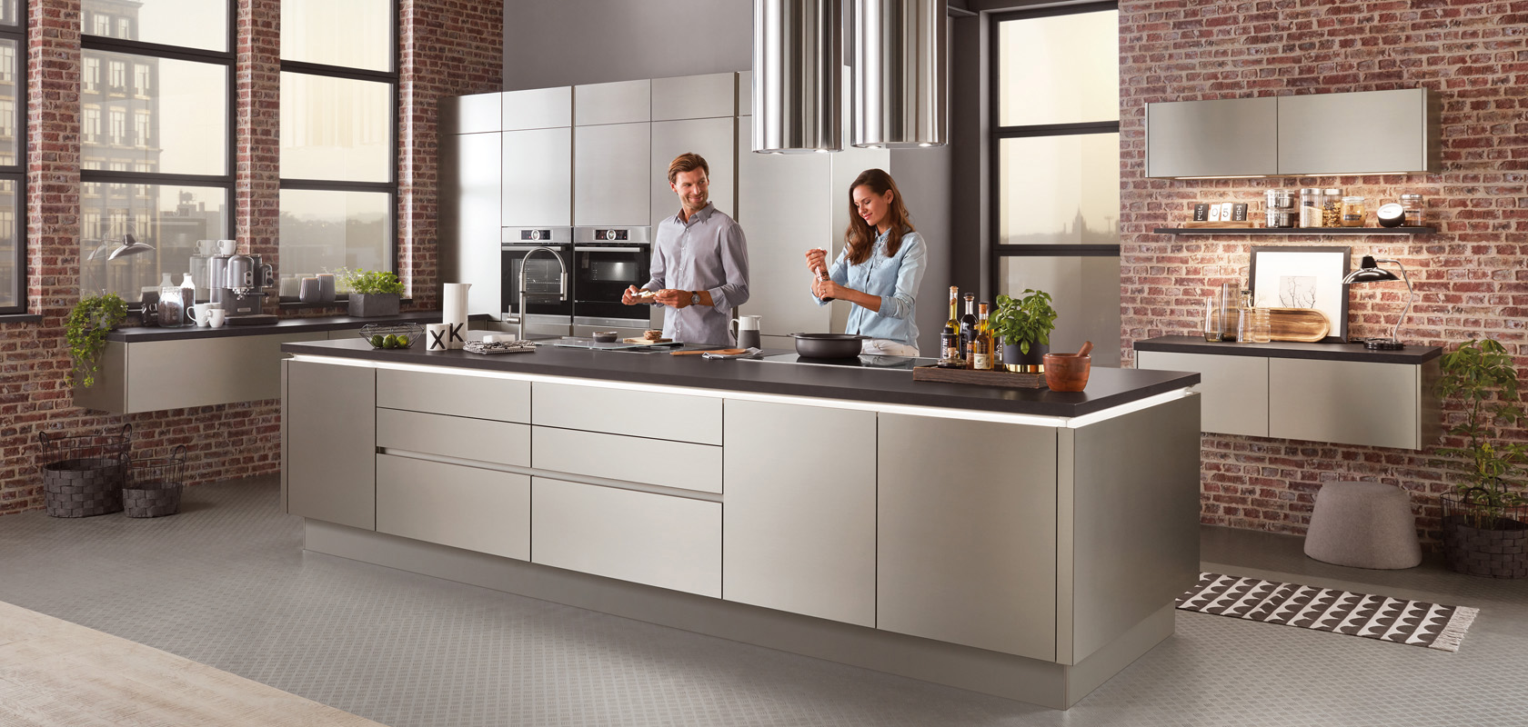 Moderní kuchyňské prostředí s párem vařícím společně, s nerezovými spotřebiči, elegantními skříňkami a průmyslově stylovou cihlovou stěnou v pozadí.