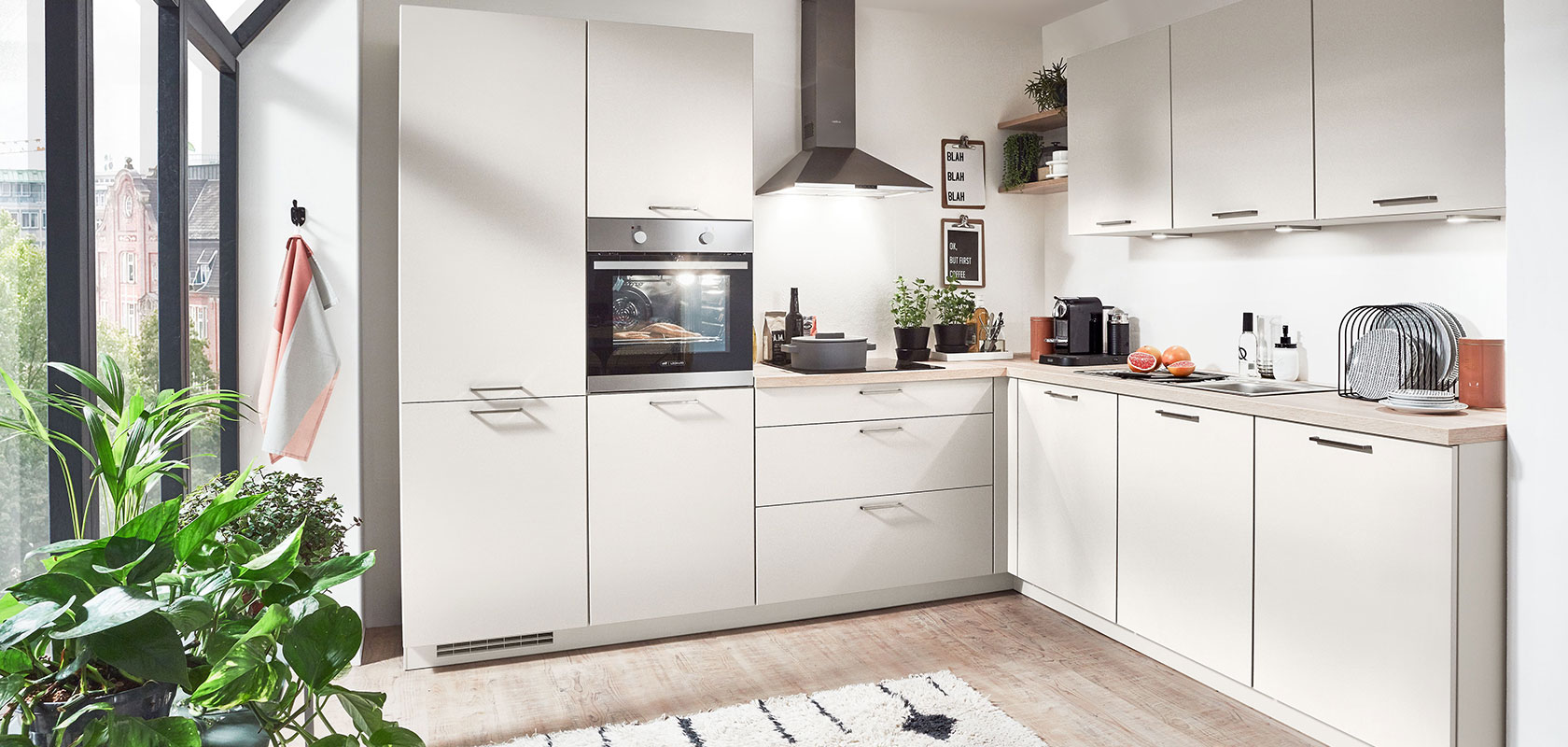 Interni di cucina moderna con armadi bianchi, elettrodomestici integrati e un tocco di verde vicino alla finestra, che illustra un design urbano pulito e contemporaneo.