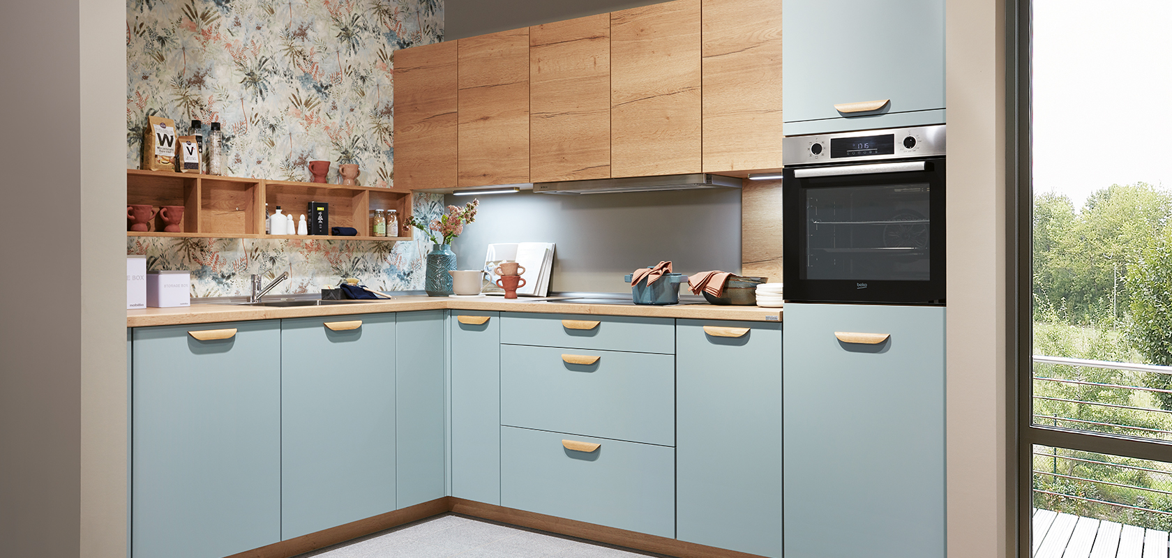Moderne Kücheninnenräume mit blauen Schränken, Holzarbeitsplatten und Edelstahlgeräten, akzentuiert mit Blumenmuster-Tapete und natürlichem Licht von einem Fenster.