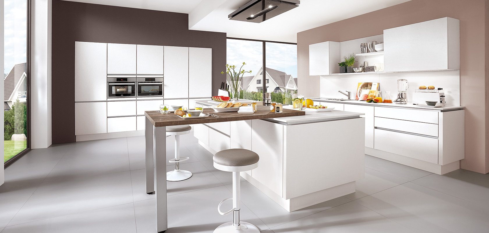 Interior de cocina moderna con encimeras blancas limpias, electrodomésticos integrados elegantes y una barra de desayuno situada frente a grandes ventanas con vistas a un entorno suburbano.