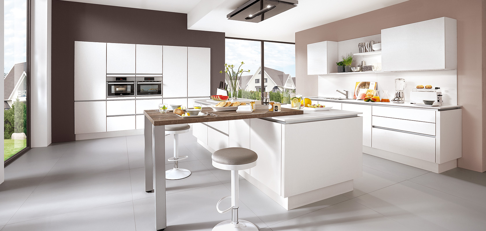 Modern kitchen interior with clean white countertops, sleek built-in appliances, and a breakfast bar set against a backdrop of large windows with a suburban view.