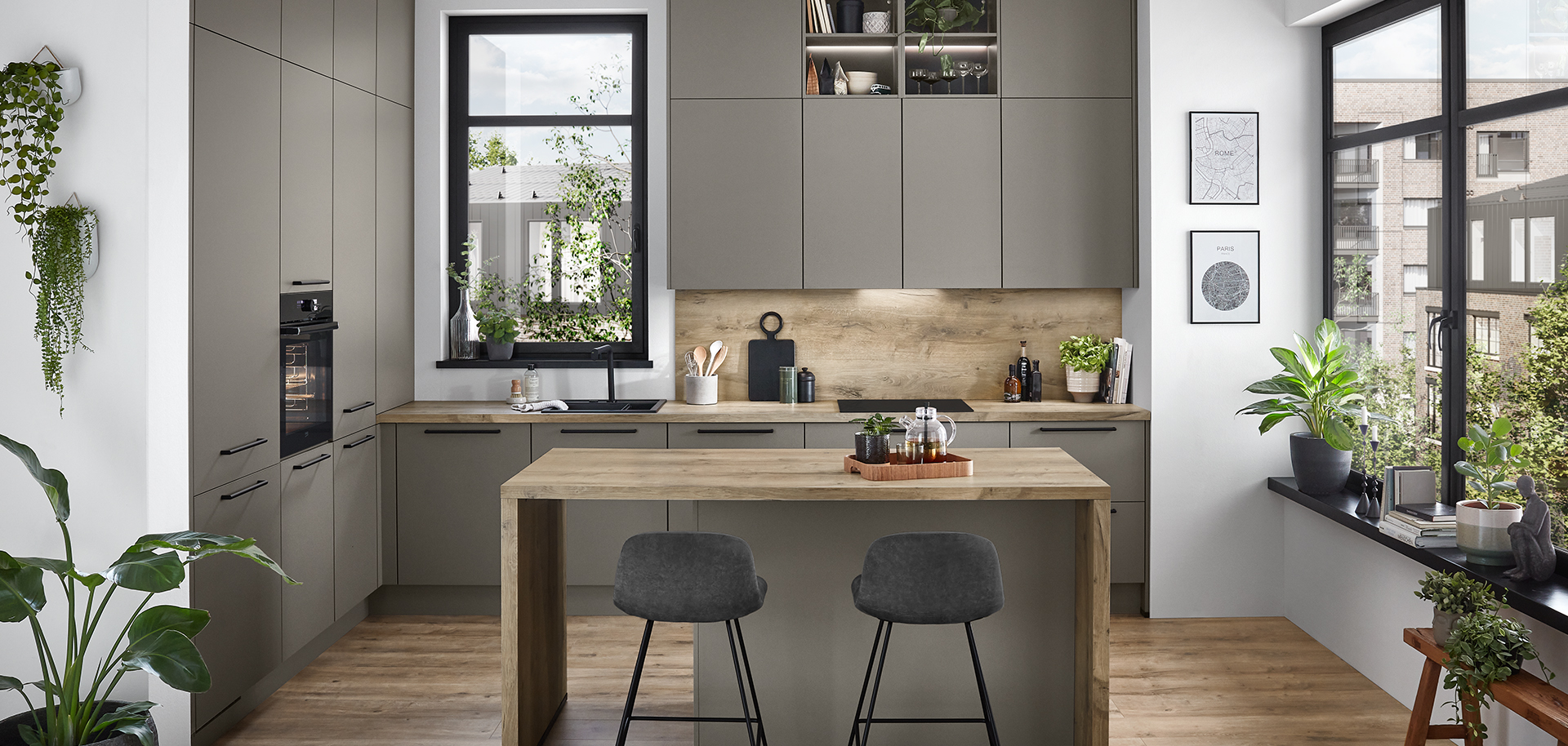Diseño de cocina moderna con elegantes gabinetes grises, pisos de madera, una isla central con taburetes, y luz natural complementada por plantas de interior.