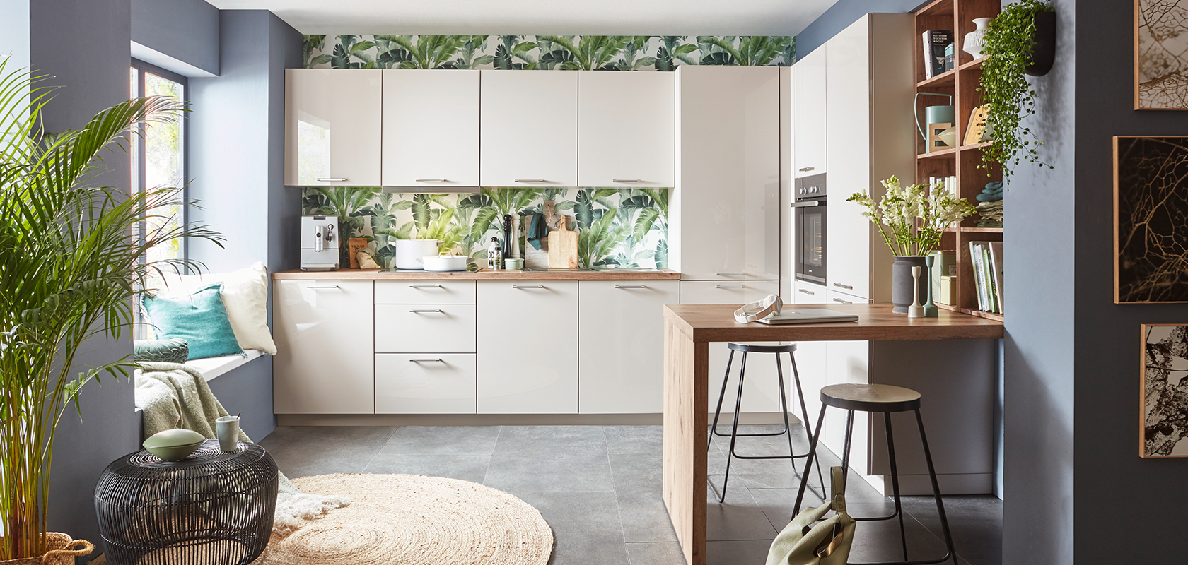 Intérieur de cuisine moderne avec des armoires blanches, un papier peint tropical, un comptoir en bois, des tabourets de bar élégants et des accents de plantes vertes créant un espace confortable et contemporain.