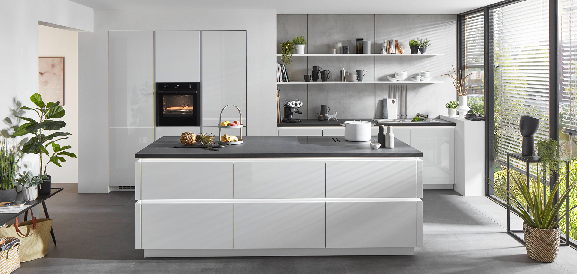 Cocina moderna con gabinetes blancos, electrodomésticos integrados y una isla central con una encimera oscura, complementada por luz natural y decoración minimalista.