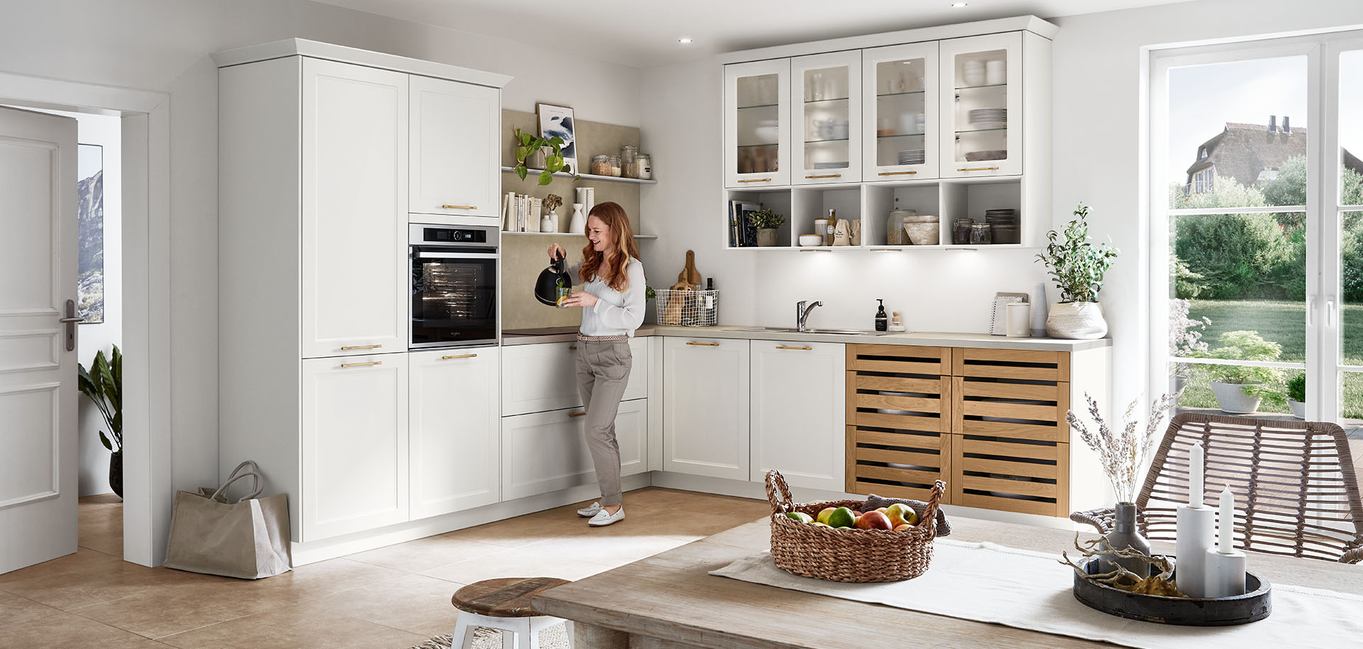 Una cocina brillante y moderna con gabinetes blancos y acentos de madera. Una persona está de pie, interactuando con un dispositivo inteligente, en medio del espacio lleno de luz solar.