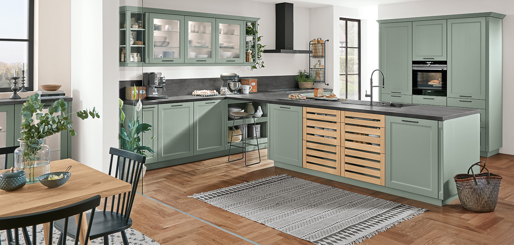Geräumige Küche mit salbeigrünen Schränken, modernen Geräten und einem Holzboden, akzentuiert von natürlichem Licht und frischem Grün.