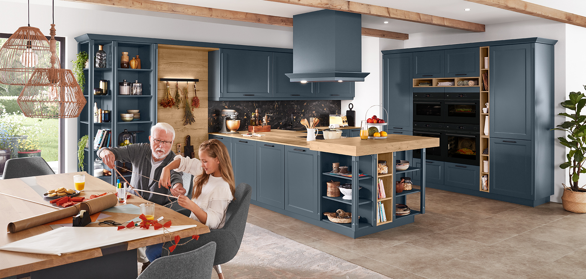 A spacious, modern kitchen with navy blue cabinetry, stainless steel appliances, and an elder crafting with a child at a sunlit wooden table.