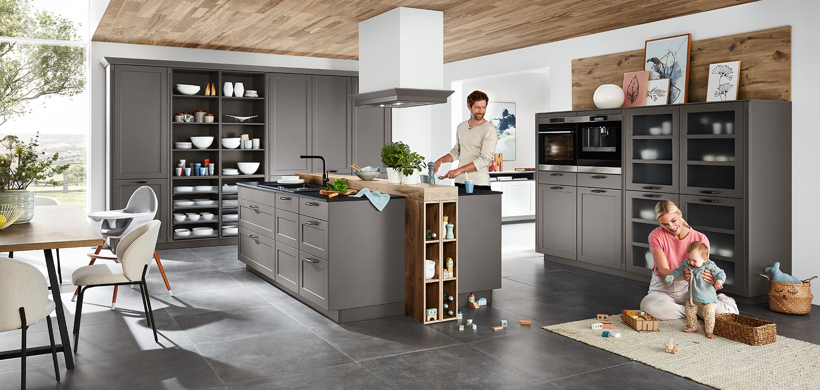 Escena de cocina contemporánea con una familia; un adulto cocinando, y otro jugando con un niño entre gabinetes grises elegantes y electrodomésticos modernos.