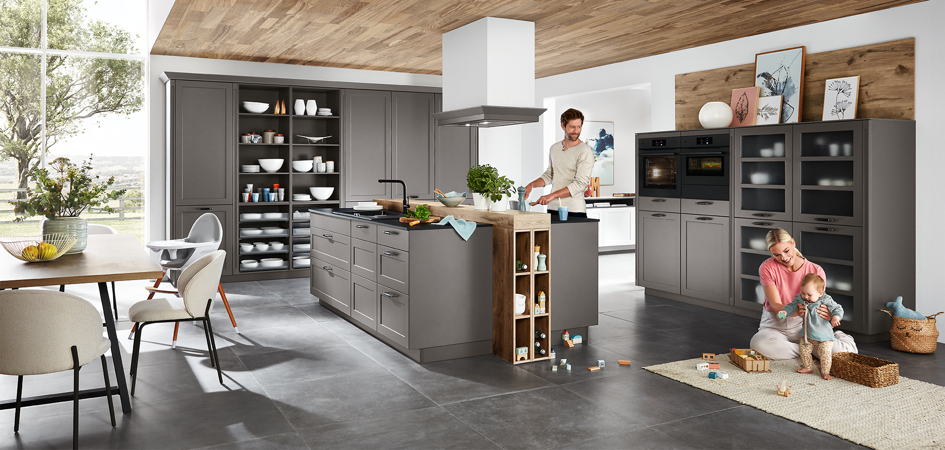 Contemporary kitchen scene with a family; an adult cooking, and another playing with a child amidst stylish gray cabinetry and modern appliances.