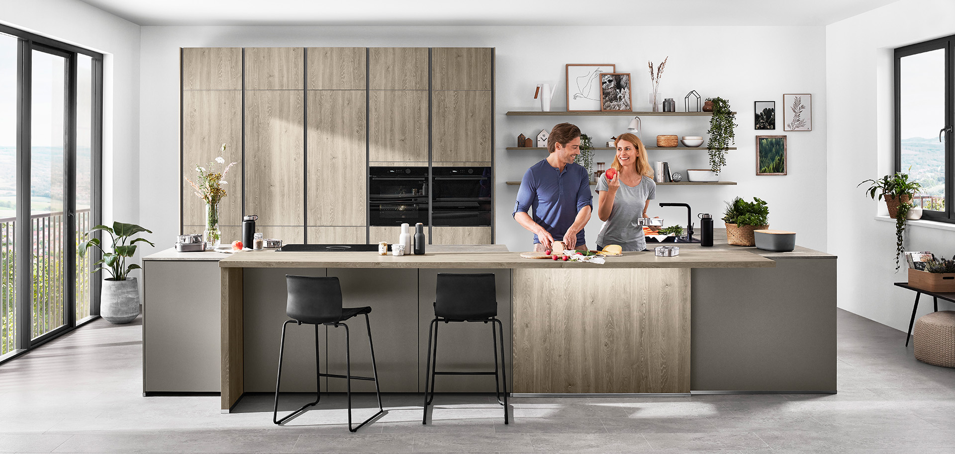 Una cocina moderna con una pareja preparando comida juntos, con gabinetes elegantes, electrodomésticos integrados y una isla con taburetes que da a una vista panorámica.
