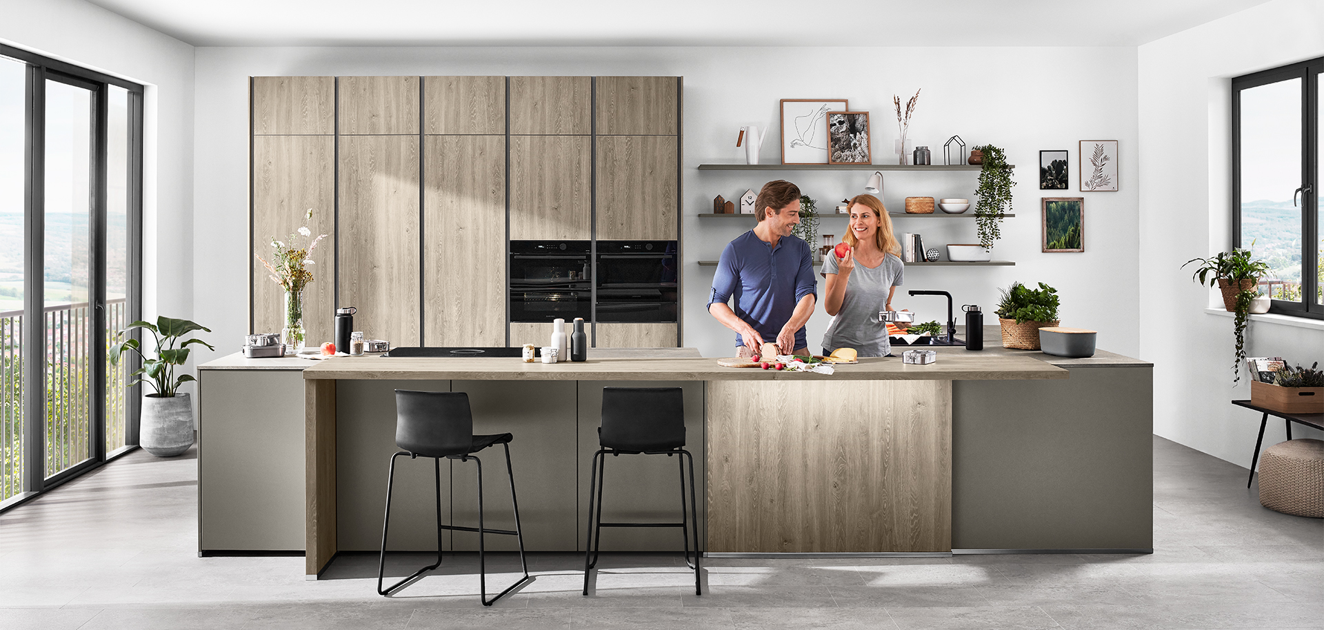 A modern kitchen with a couple preparing food together, featuring sleek cabinetry, integrated appliances, and an island with bar stools overlooking a scenic view.