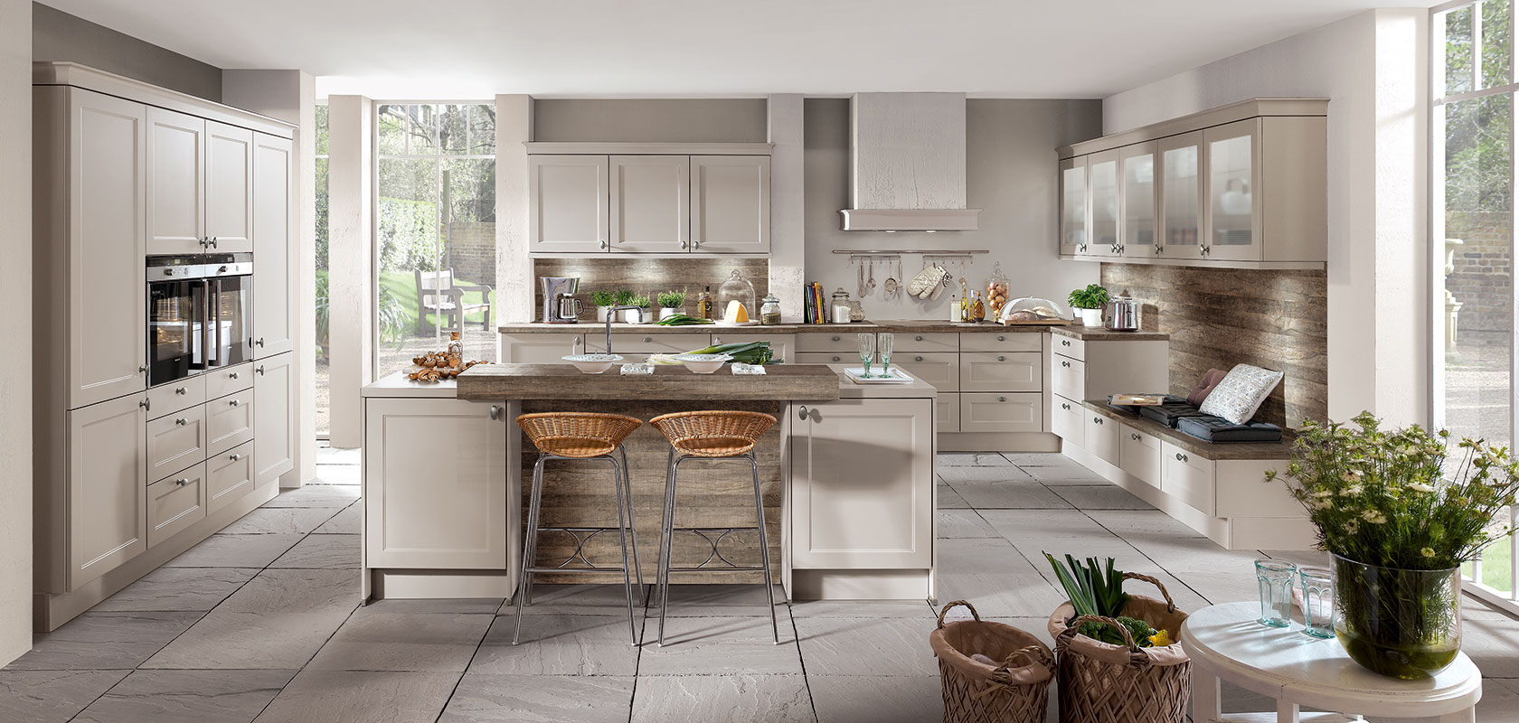 Espaciosa cocina moderna con gabinetes beige, una isla con taburetes de mimbre y electrodomésticos de acero inoxidable iluminados por luz natural de grandes ventanas.