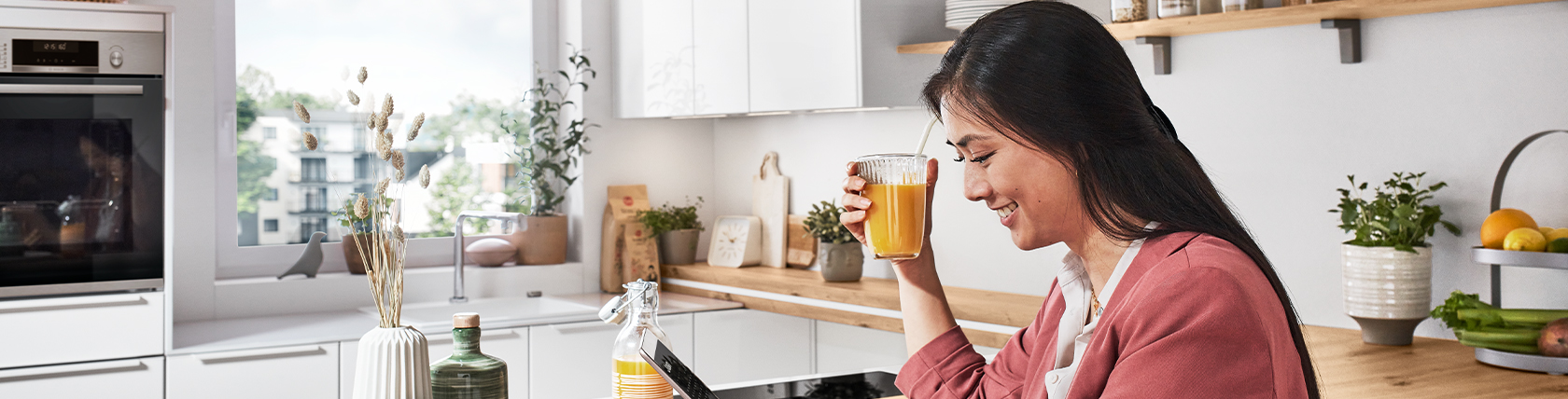 Eine lächelnde Frau genießt ein Glas Saft in einer sonnendurchfluteten, modernen Küche, die mit grünen Pflanzen und frischen Produkten gefüllt ist.