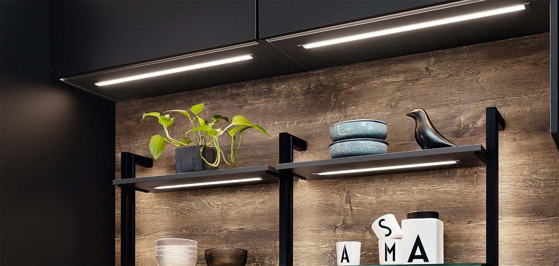 Detailaufnahme einer L-Küche mit dunkler Oberschrankbeleuchtung, offenen Regalen vor Holzwand und dekorativen Elementen wie Keramik, Pflanze und Vogelfigur.