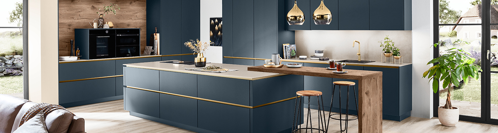 Contemporary kitchen with navy blue cabinetry, brass accents, integrated appliances, and a wooden breakfast bar, complemented by natural light and greenery.