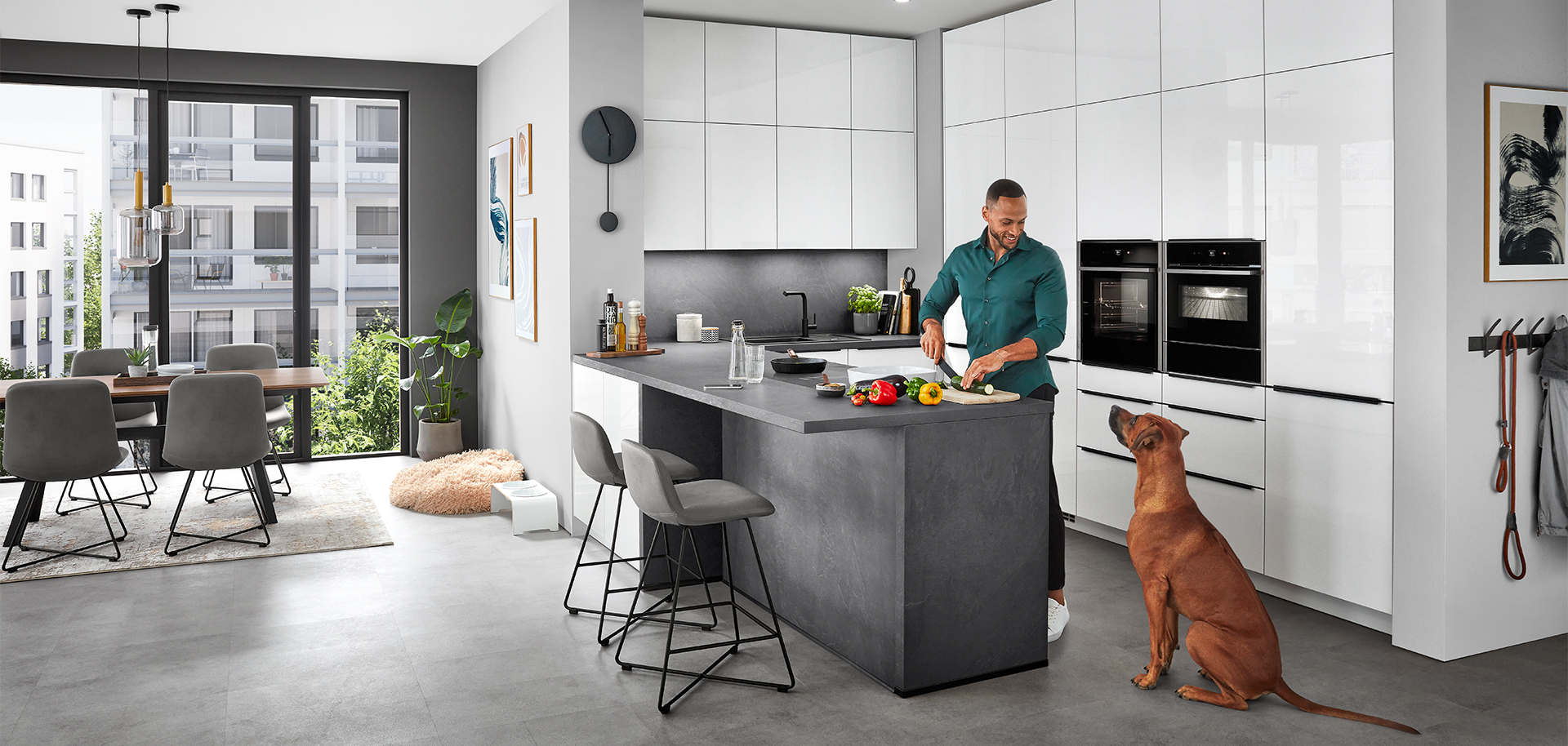 Stylowa nowoczesna scena kuchenna z mężczyzną przygotowującym jedzenie na wyspie kuchennej, przyglądającym się pieskiem, odzwierciedlająca wygodny, współczesny styl życia w domu.