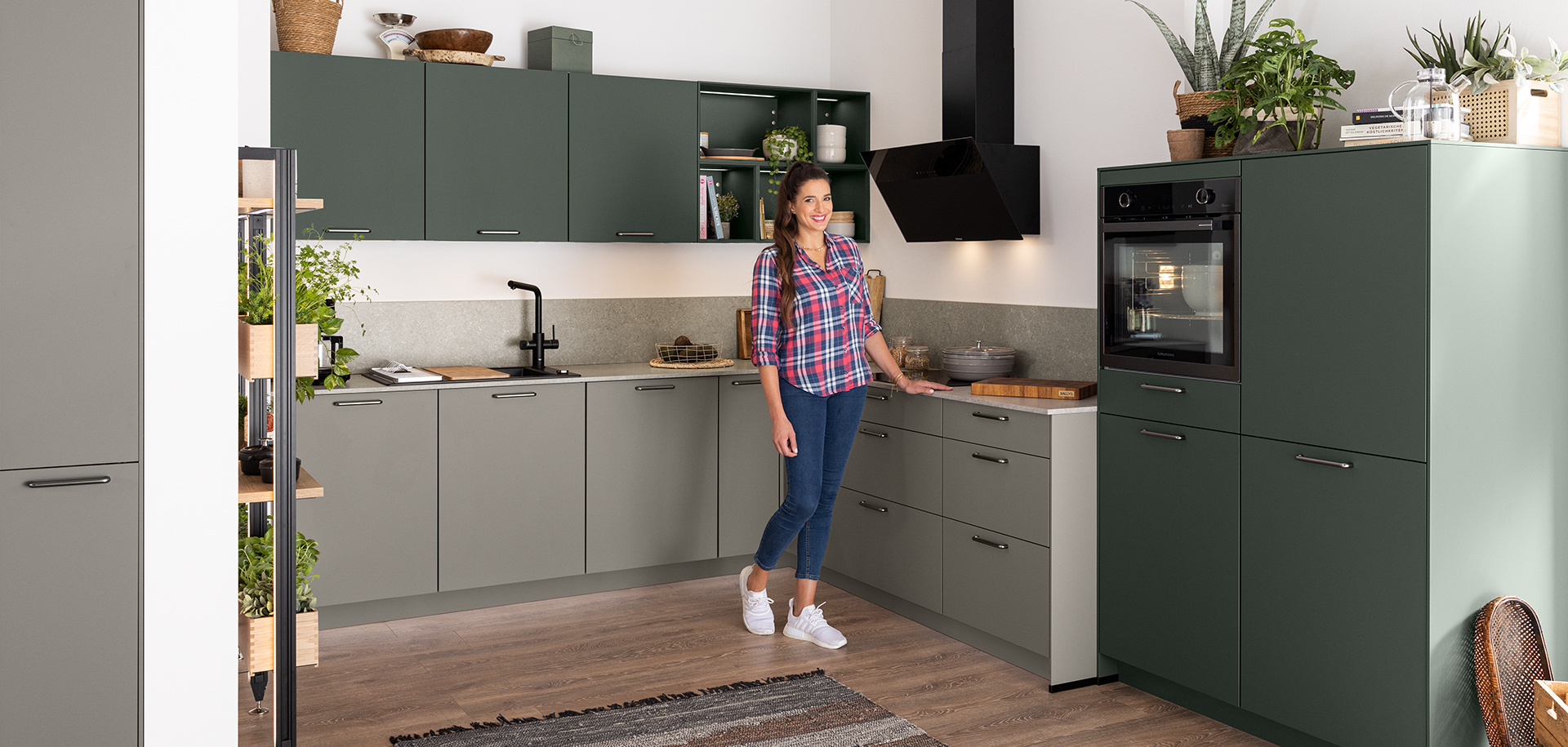 Žena stojící v moderní kuchyni s zelenými skříňkami, nerezovými spotřebiči a dřevěnými doplňky, dávající přívětivý gesto.