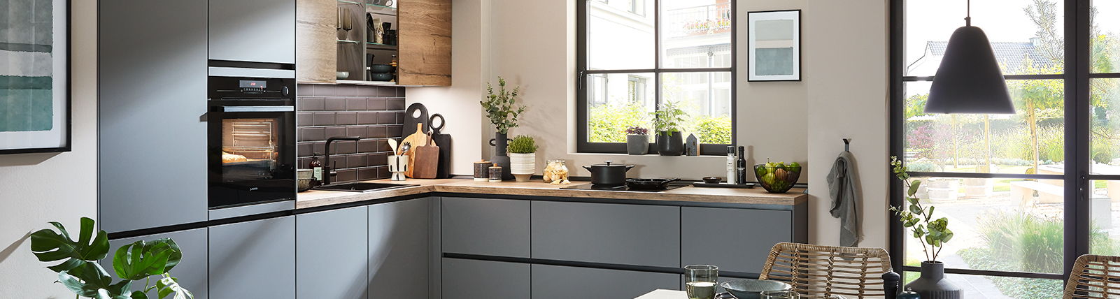 Moderne L-Küche mit matten Fronten in Grauton und warmen Holzelementen, kombiniert mit schwarzen Akzenten und großen Fensterflächen für ein helles Raumgefühl.