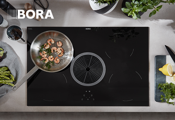 Moderna placa de inducción BORA con un sistema de extracción integrado, mostrando un diseño elegante y una sartén con camarones cocinándose, rodeada de ingredientes frescos.